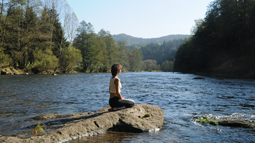 Eine Frau sitzt im Schneidersitz auf einem Felsen mitten im Fluss, ringsum bewaldete Hänge