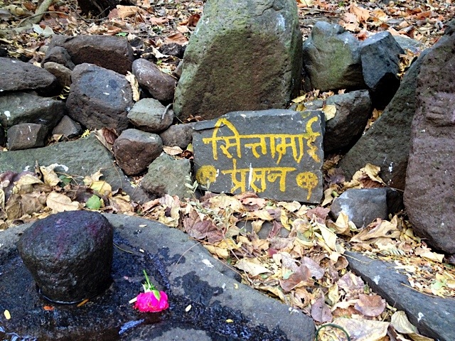 Ein kleiner Schrein aus geschichteten Steinen im Wald, mit beschrifteter Tafel, einem Lingam und einer roten Rose auf dem Laub