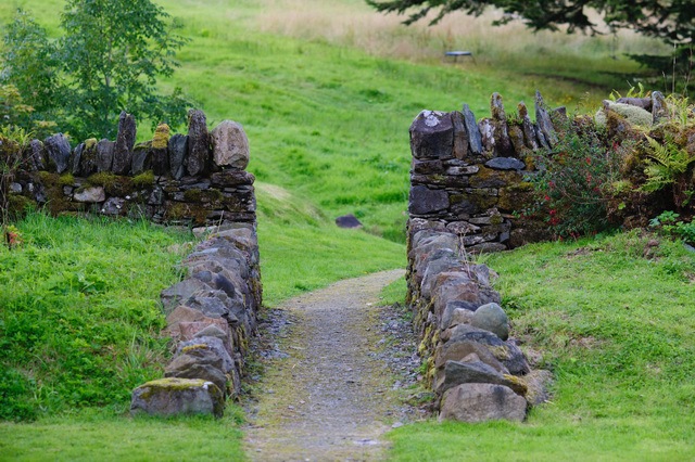 Ein schmaler Weg führt über eine alte Steinbrücke zwischen Trockenmauern eine grüne Wiese hinauf